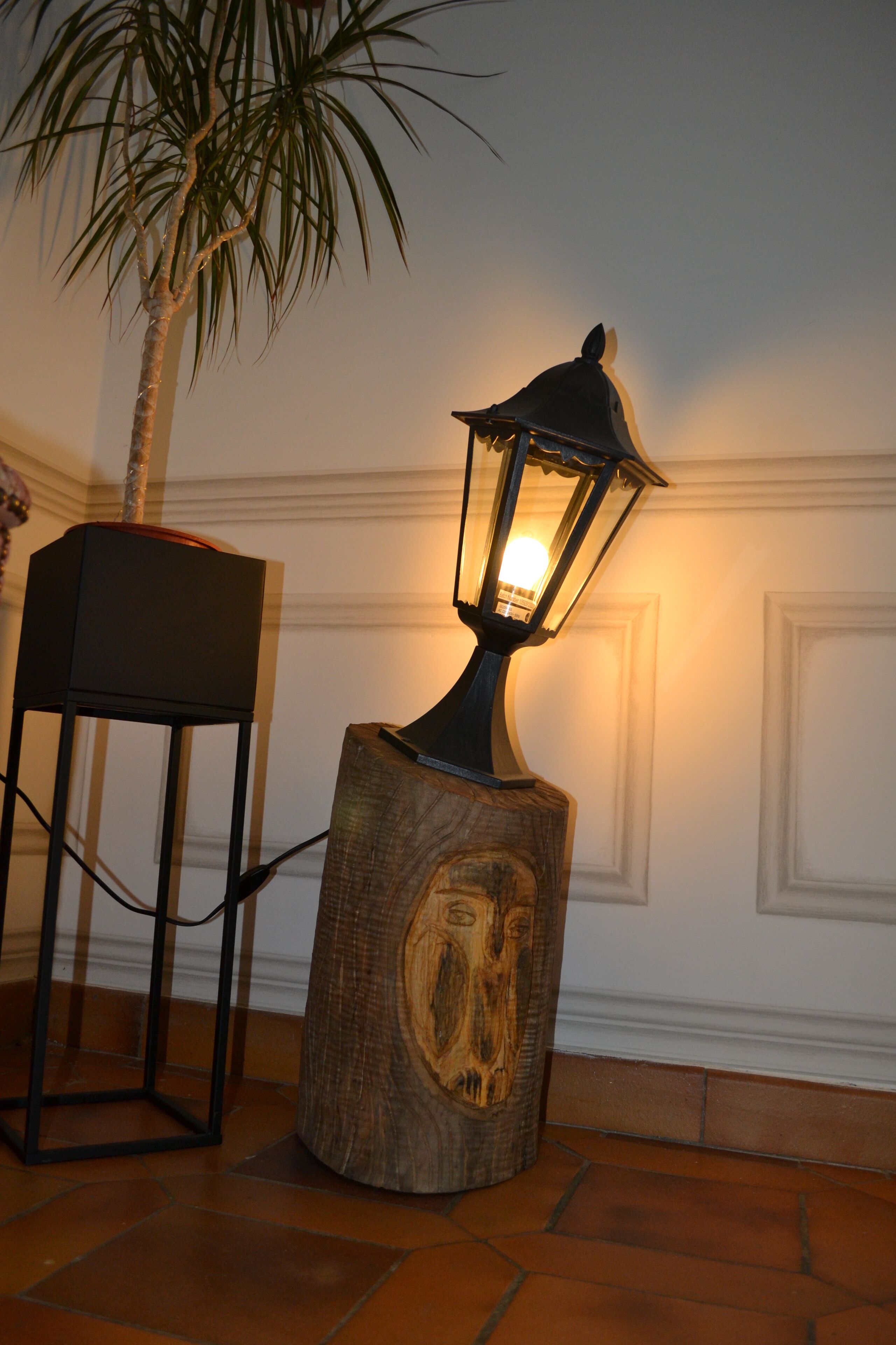 A wooden lamp with a carved design on a pedestal, a small table with a lamp, and a palm tree in the background.
