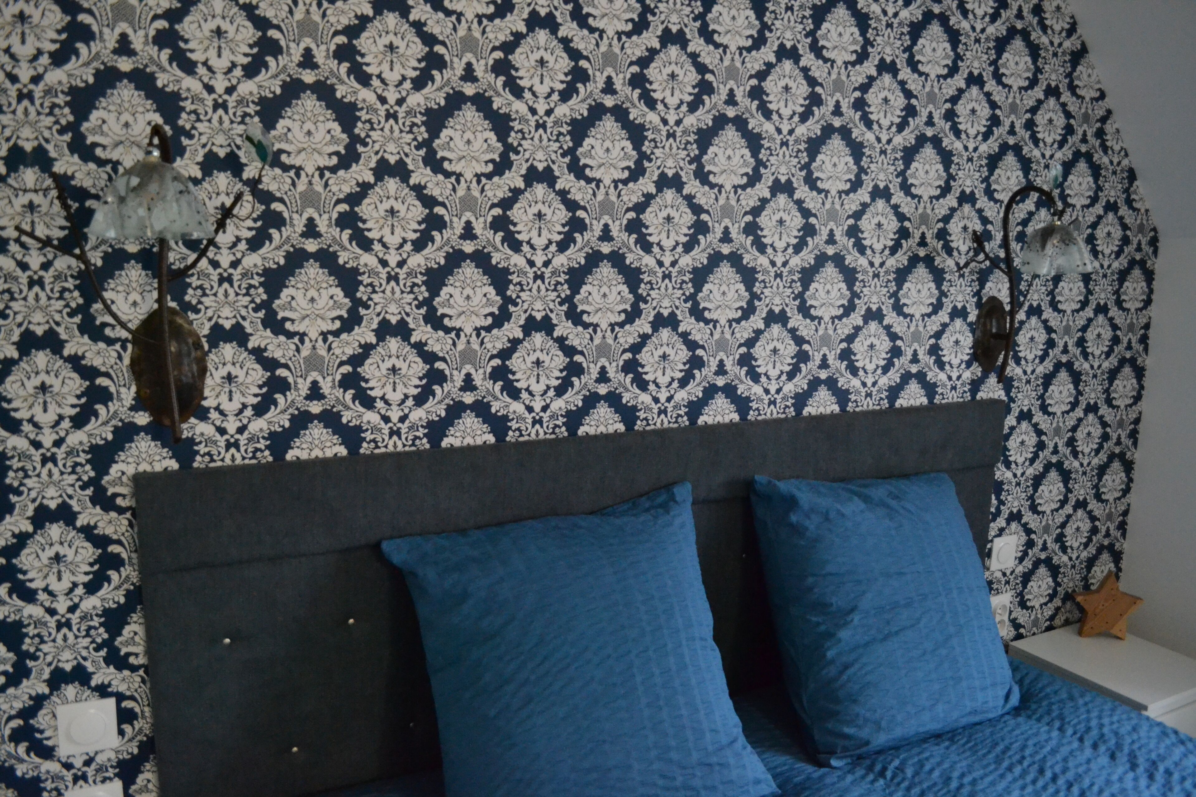 A bedroom with a patterned wallpaper, a grey headboard, and two blue pillows.