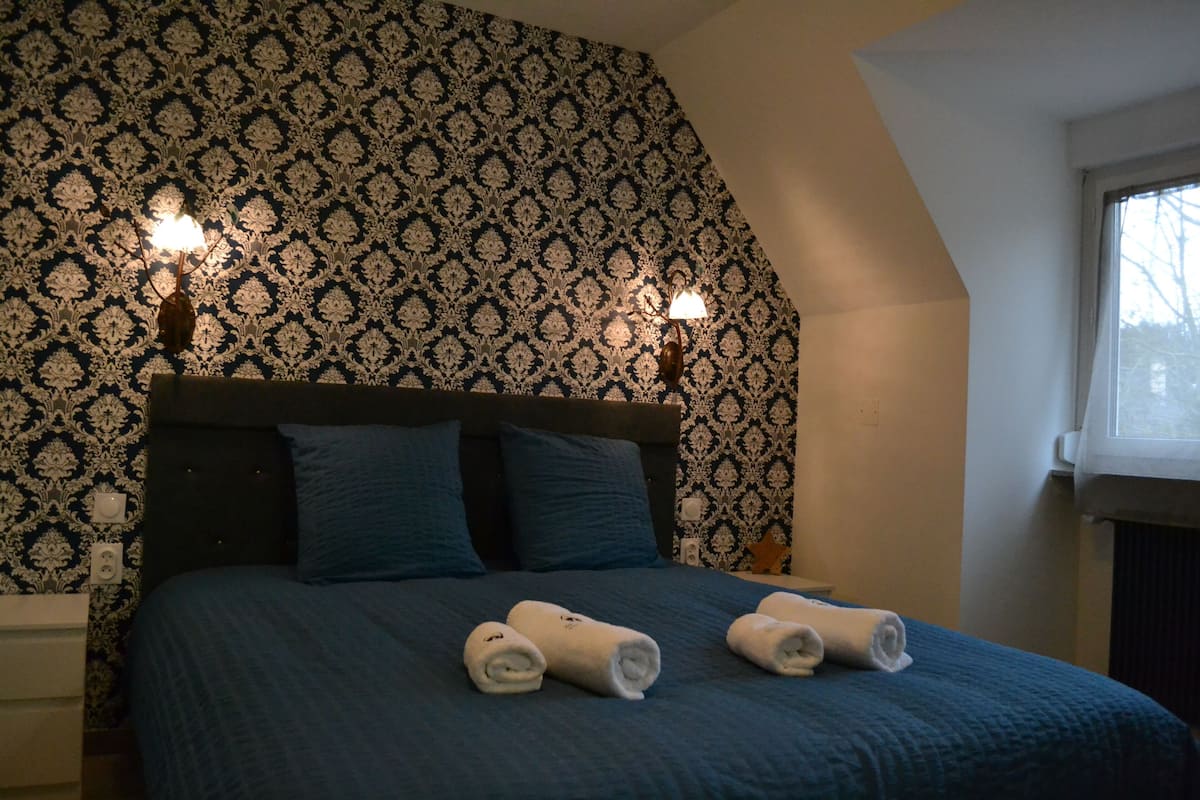 Une chambre avec un papier peint à motifs, une tête de lit sombre, deux coussins bleus et deux serviettes enroulées posées sur le lit.