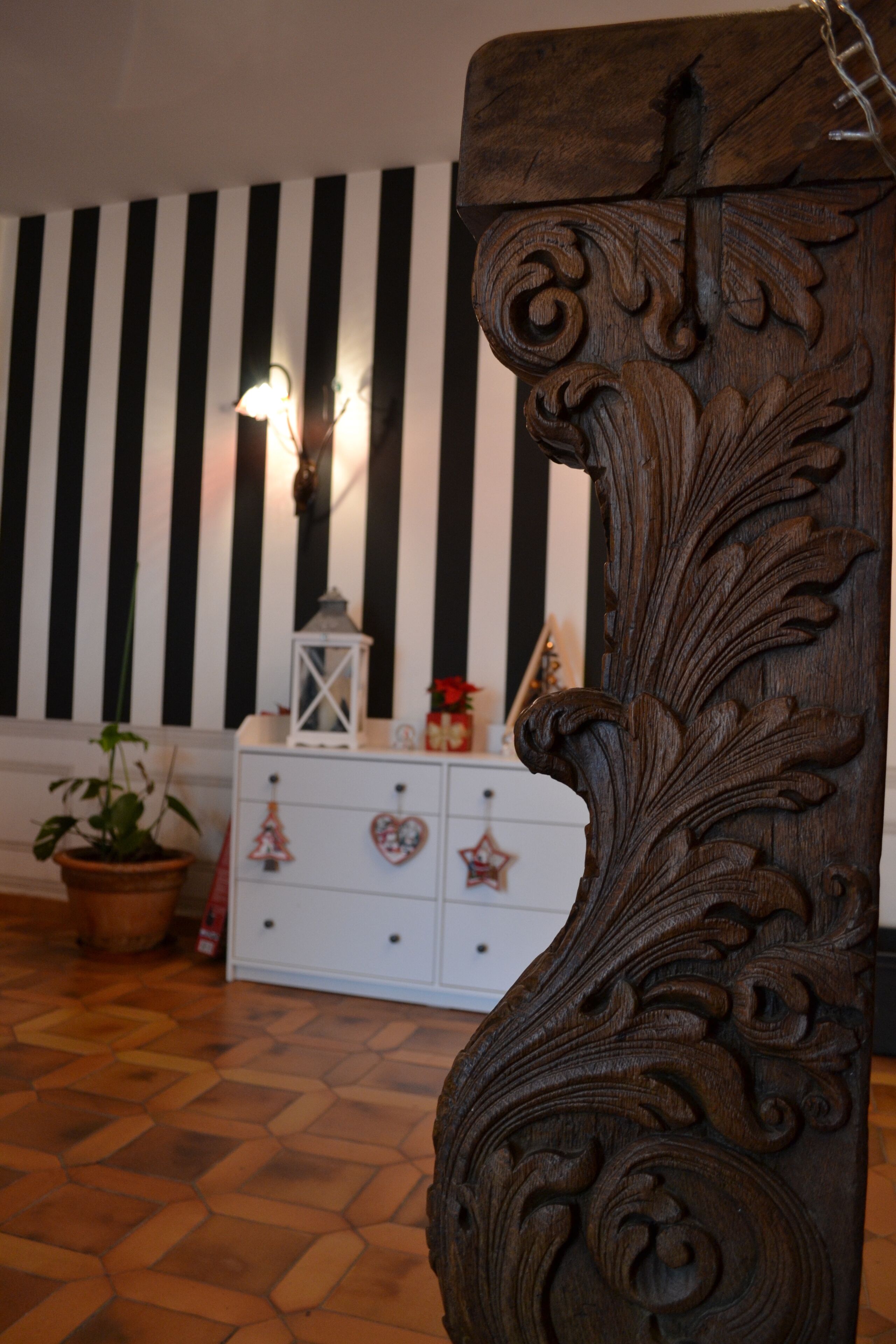 A wooden carved chair with intricate designs, a white dresser with heart-shaped ornaments, a potted plant, and a striped wall in the background.