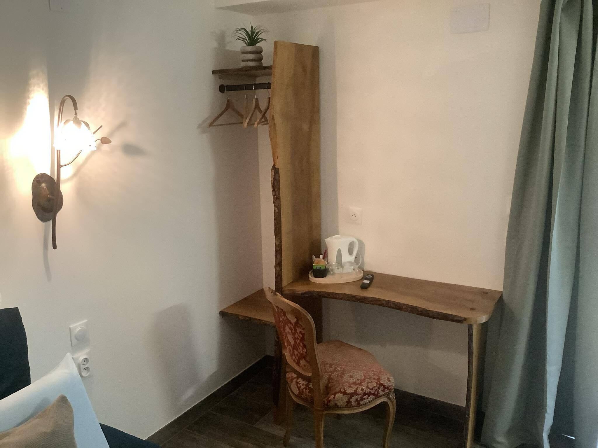 A corner room with a wooden desk, a chair, a wall-mounted light, a plant, and a small shelf.