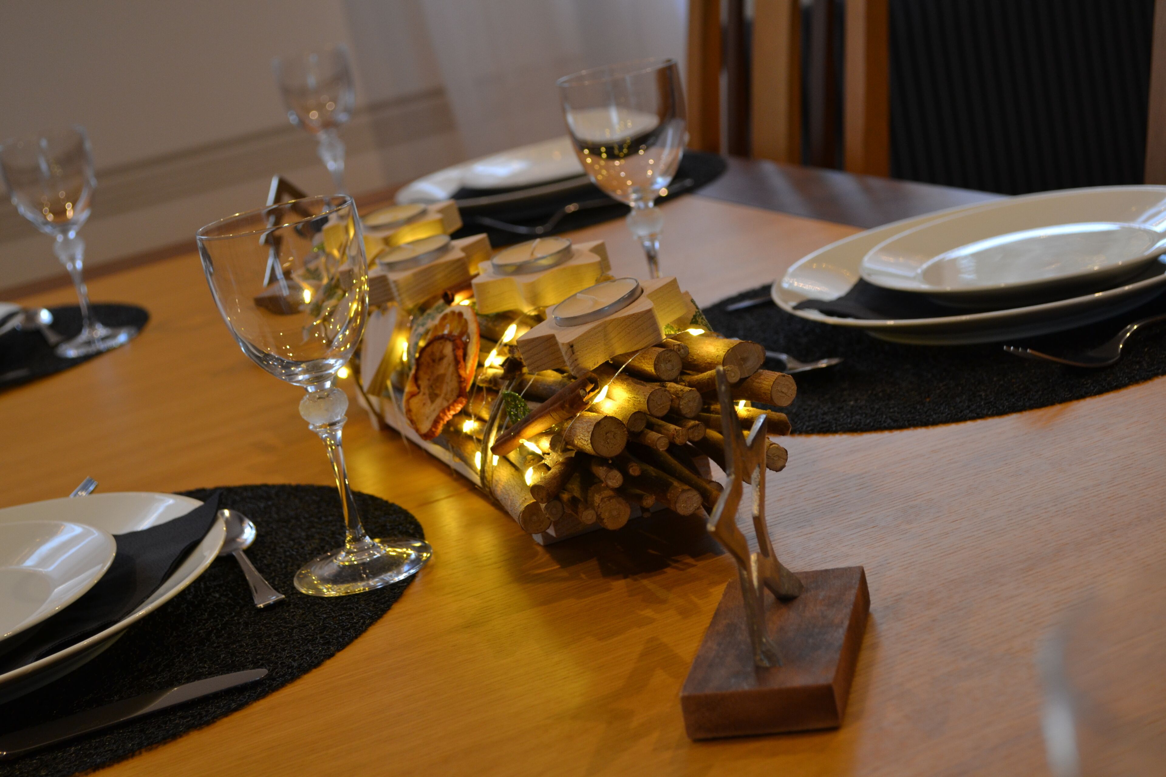 Una mesa de comedor dispuesta con platos, cubiertos y copas de vino, con un centro decorativo de velas y leña.
