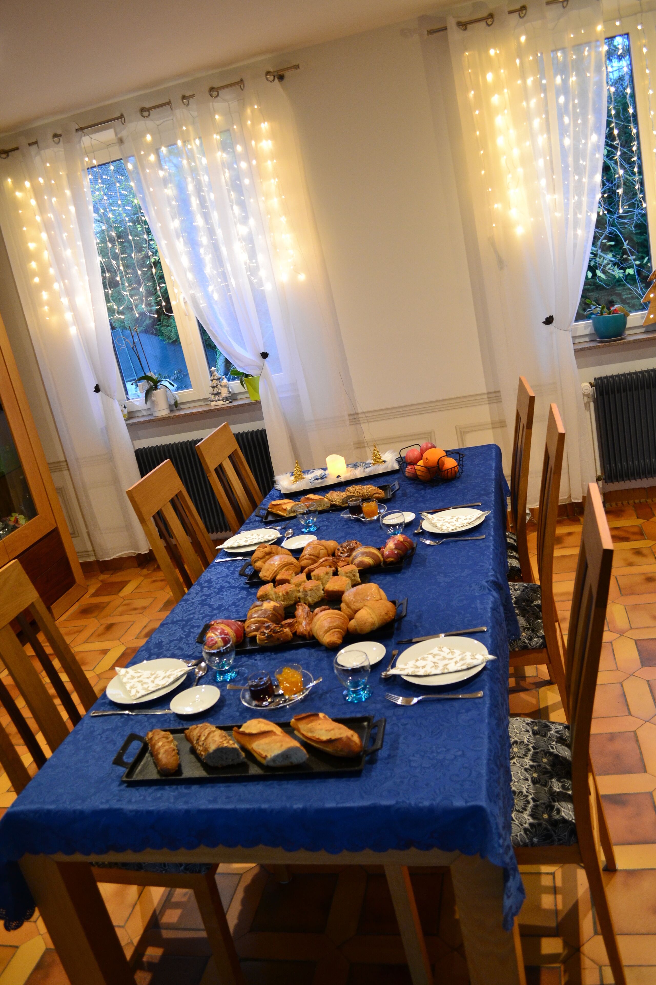 Mesa de comedor preparada con comida y bebidas, rodeada de sillas, cortinas y luces colgantes.