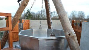 Un grand bain à remous en métal, suspendu par des chaînes, entouré de structures en bois et d’un mur en béton.