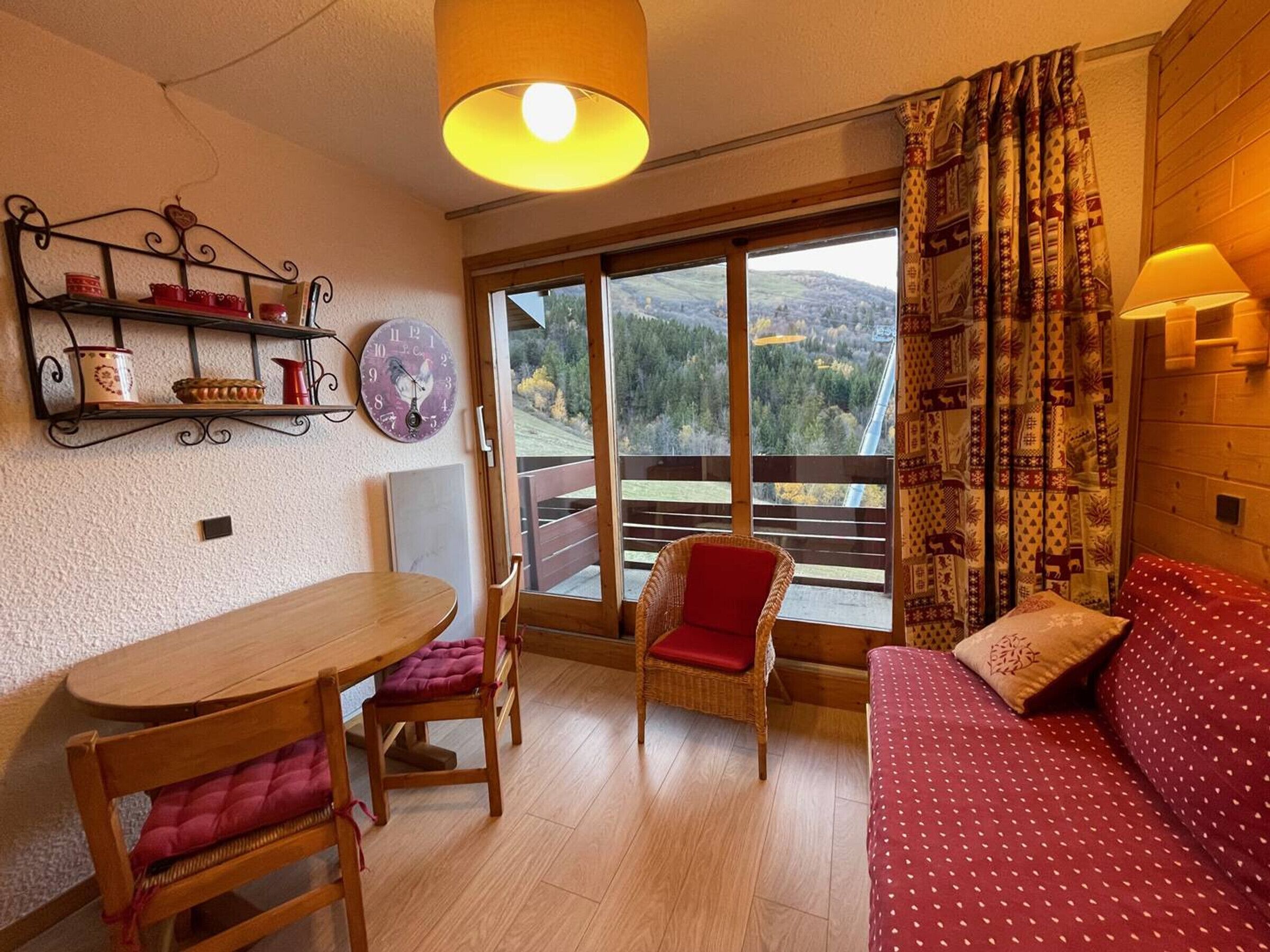 Une pièce chaleureuse avec une table en bois, deux chaises, un canapé, une grande fenêtre donnant sur des collines et une étagère murale agrémentée d’objets décoratifs.