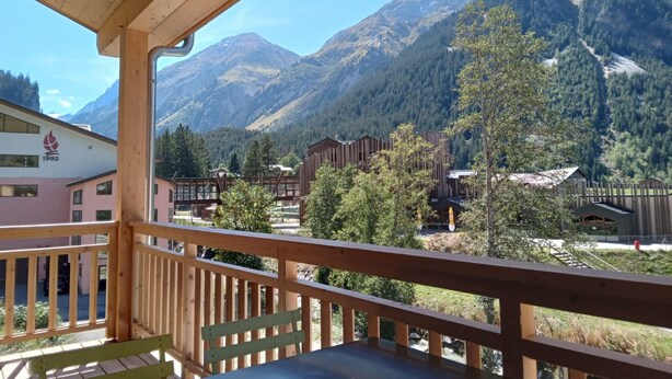 Un balcon en bois avec une table et des chaises, donnant sur un paysage montagneux avec des bâtiments en arrière-plan.