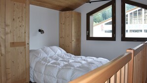 Une chambre avec un plafond en bois, un lit recouvert d’une couette blanche et deux fenêtres donnant sur un immeuble et des arbres.