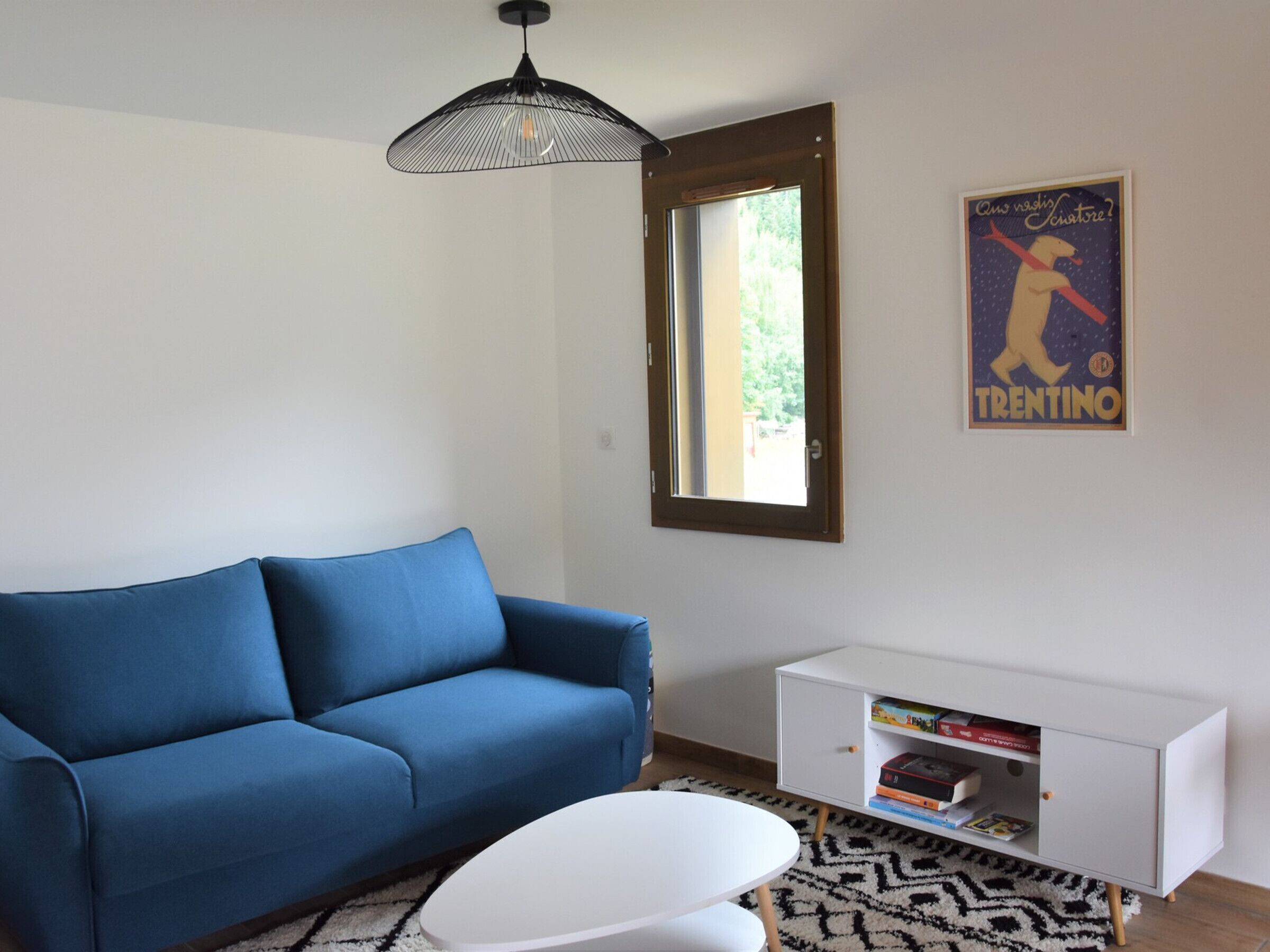 A modern living room with a blue sofa, a white coffee table, a white sideboard with books, a black and white rug, and a window with a view of greenery.