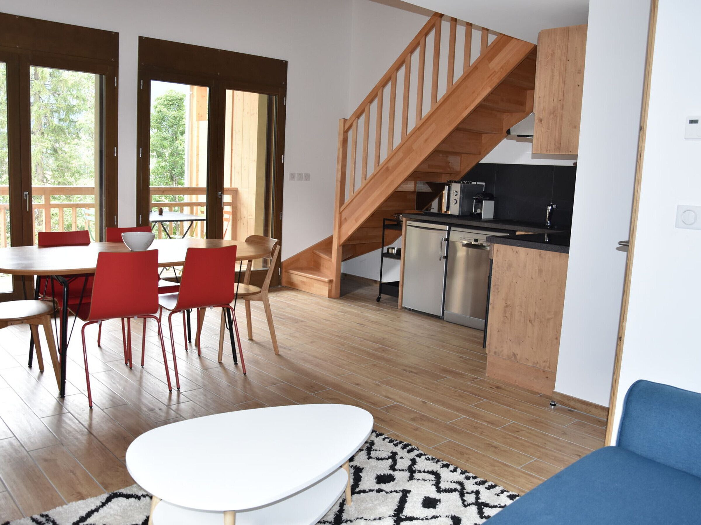 A modern kitchen with a wooden staircase, a dining area with red chairs, and a large window.