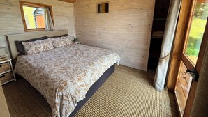 Ein Schlafzimmer mit Holzwänden, einem Bett, einem Nachttisch, einem kleinen Fenster und Blick ins Freie.