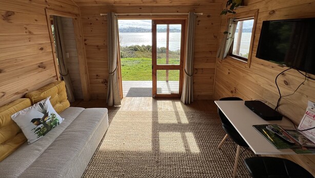 Ein Zimmer in einer Holzhütte mit einer Couch, einem Tisch, einem Fernseher und Blick auf einen See.
