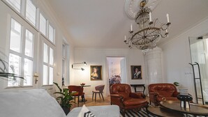 A spacious living room with large windows, a chandelier, leather armchairs, and a patterned floor.