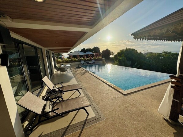 Un área de piscina moderna al aire libre con barandilla de vidrio, sillas de descanso y vista al atardecer sobre el agua.