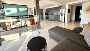 Ein modernes Wohnzimmer mit einem großen Flachbildfernseher, einem grauen Sofa und einem Essbereich mit einer Küche im Hintergrund.