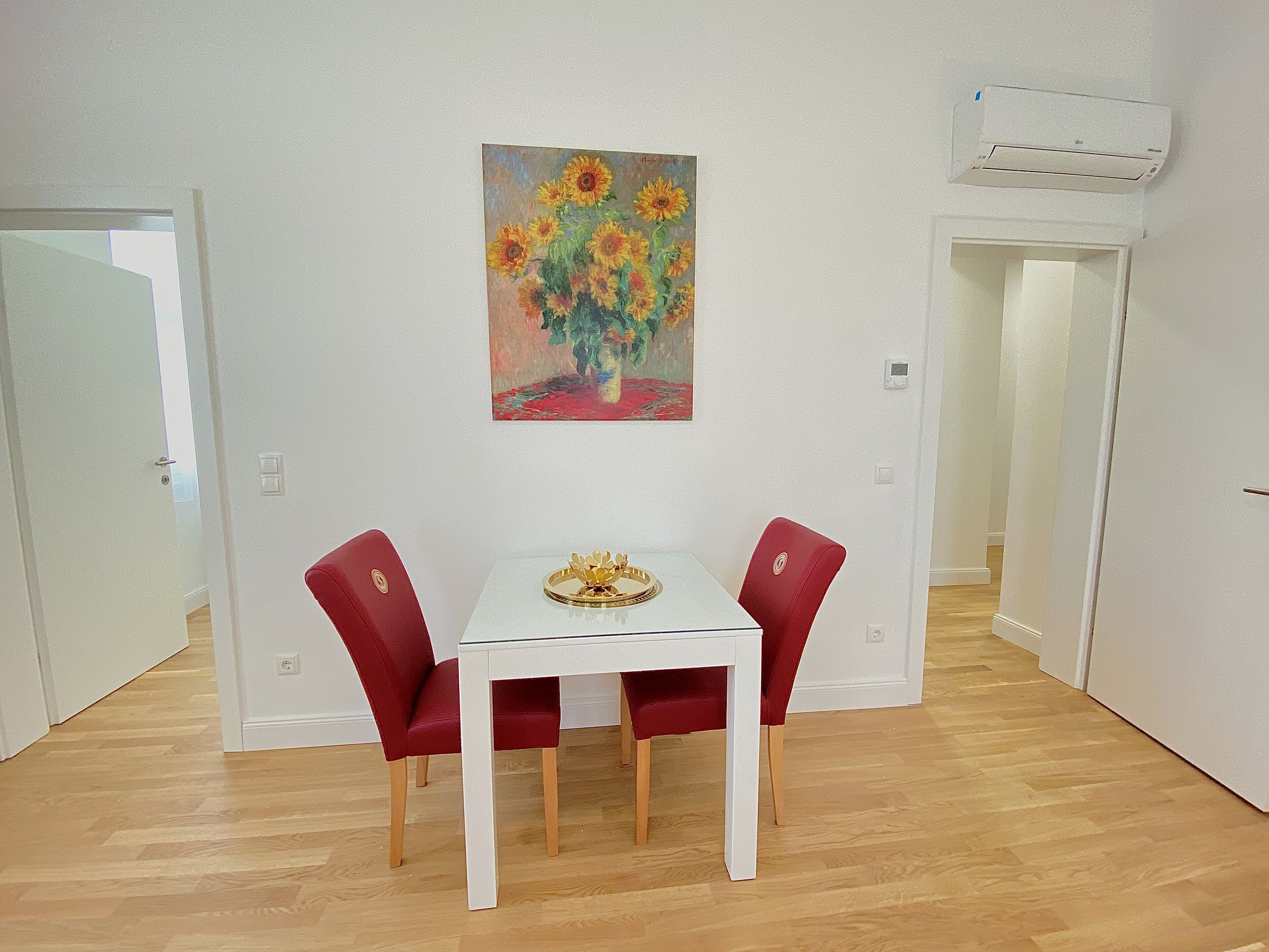 A dining area with a white table and two red chairs, a painting of sunflowers on the wall, and an air conditioning unit above.