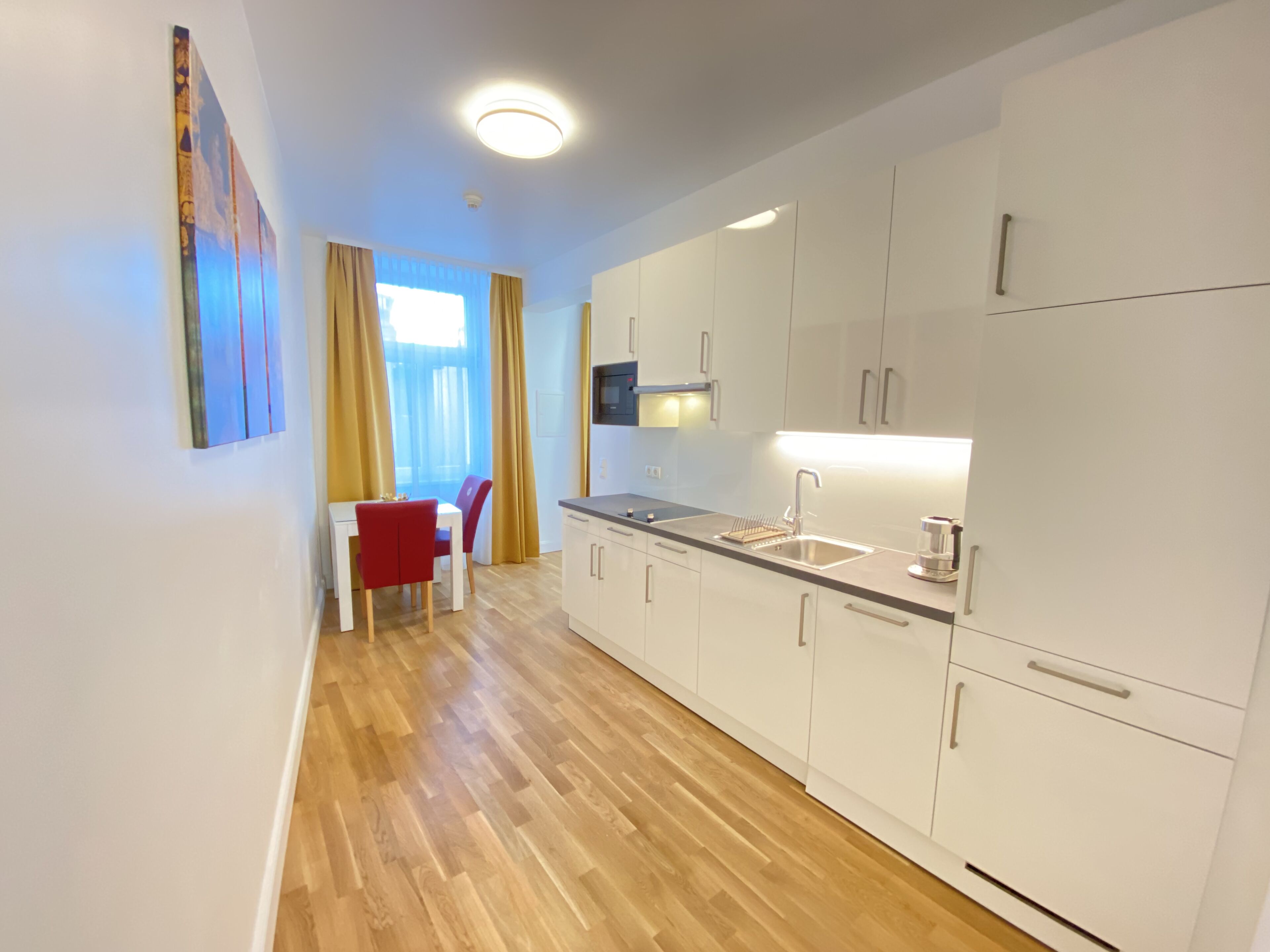 A modern kitchen with white cabinets, a stainless steel sink, and a microwave. There is a dining area with a red chair and a table, and a window with yellow curtains.