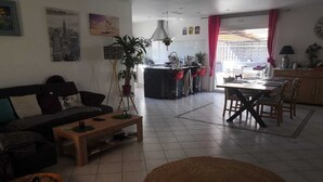 A living room with a sofa, coffee table, and a dining area with a table and chairs. There is a kitchen area with a bar and a dining table. - Maison Chaleureuse Dans le Sud (Cournonsec)