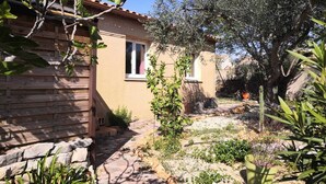A rustic stone building with a tiled roof, a small window, and a garden with various plants and a tree. - Maison Chaleureuse Dans le Sud (Cournonsec)