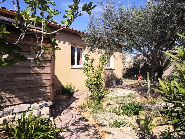 A rustic stone building with a tiled roof, a small window, and a garden with various plants and a tree.