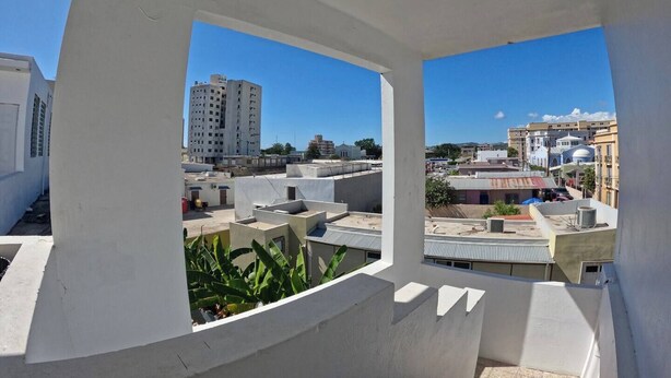 A view from a balcony with white walls, overlooking a cityscape with various buildings, including a tall structure and several smaller ones. - Apartment Double Bed- Sleeps 2 - Free WIFI (Ponce)