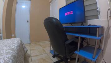 Ein Schlafzimmer mit Bett, Schreibtisch, Computer, Monitor mit der Anzeige 'NETFLIX', Stuhl und Fenster mit Jalousien.
