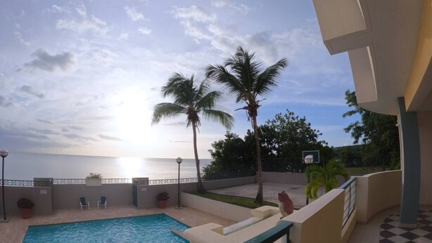 Une piscine avec vue sur le coucher de soleil sur l’océan, des palmiers et un terrain de basketball.