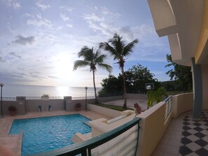 A swimming pool with a view of the sunset over the ocean, palm trees, and a basketball court.