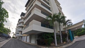 Un immeuble résidentiel à plusieurs étages, doté de balcons, d’un espace de stationnement couvert et de palmiers.