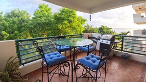 Un balcon avec une table en verre, des chaises rembourrées bleues, un barbecue et une vue sur une végétation luxuriante.