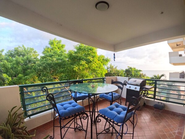 A balcony with a glass table, blue cushioned chairs, a grill, and a view of lush greenery.