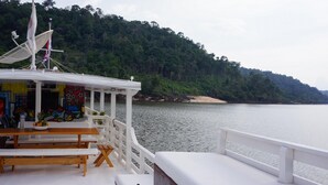 A boat with a covered seating area, a table with a bowl of fruit, and a view of a forested coastline. - Gorgeous Converted Fishing Vessel in Alter do Chao (Santarém)