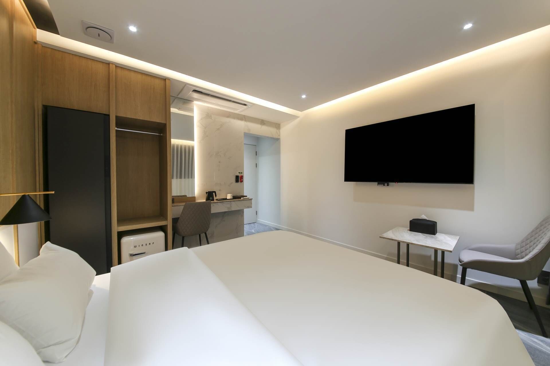 A modern hotel room with a large bed, a desk, a chair, a TV, and a wardrobe.