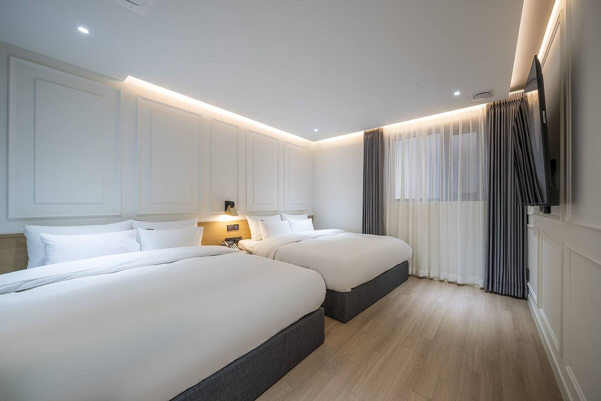 Ein Hotelzimmer mit zwei Betten, einem Flachbildfernseher, einem Nachttisch mit Lampe und einem großen Fenster mit Vorhängen.