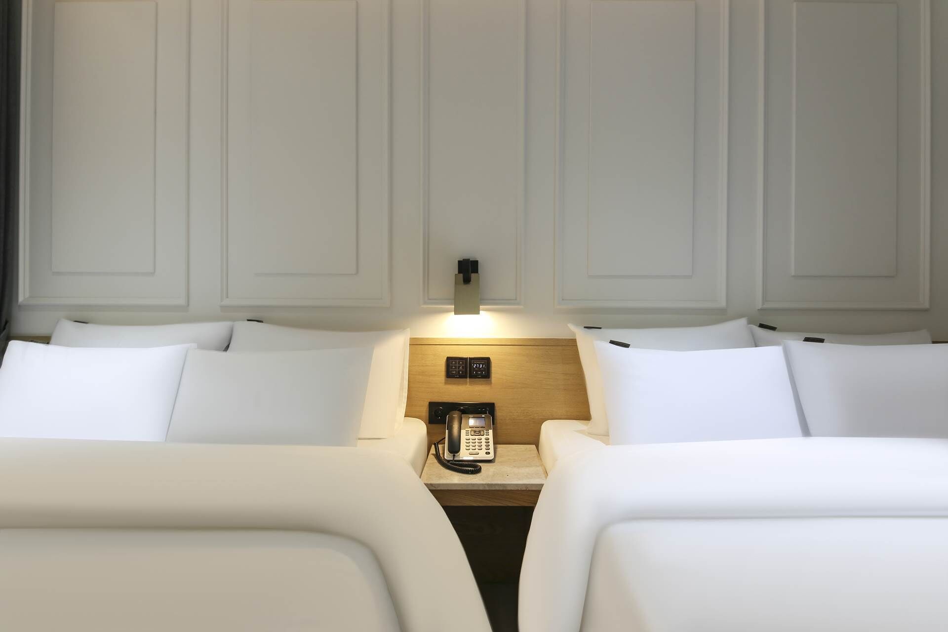 A hotel room with two beds, a bedside table with a phone, and a wall-mounted light.