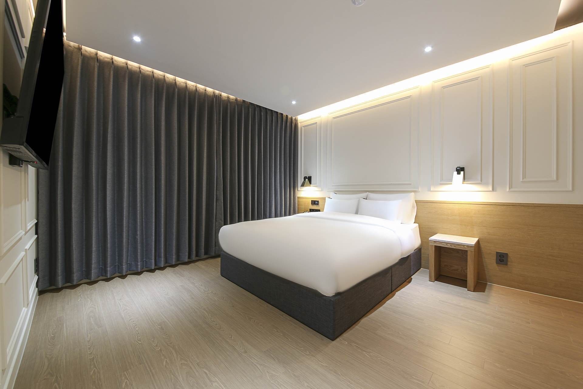 A modern hotel room with a large bed, a wooden headboard, a nightstand, a TV, and a curtain.