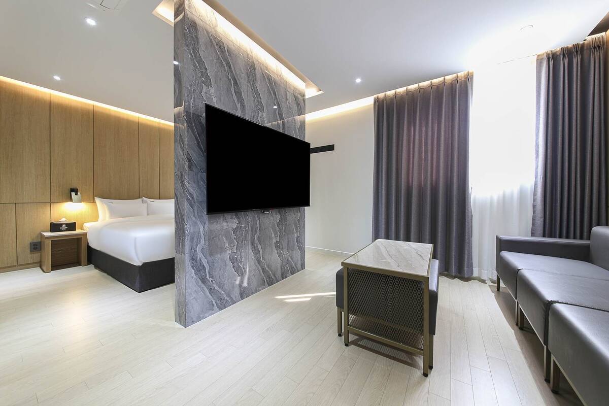 Ein modernes Hotelzimmer mit einem großen Flachbildfernseher an einer Marmorwand, einem Bett mit Holz Kopfteil, einem kleinen Tisch mit einem Netzschirm und einer Couch.