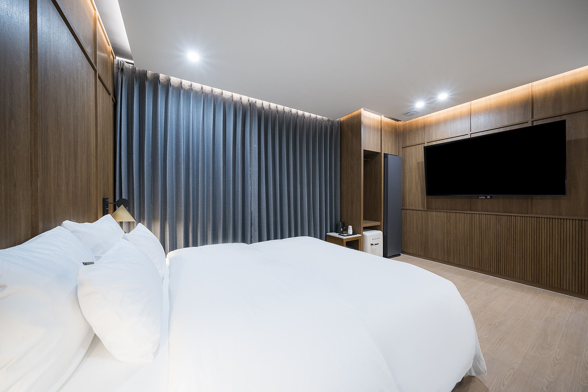 A modern hotel room with a large bed, a flat-screen TV mounted on the wall, and a small bedside table with a lamp.