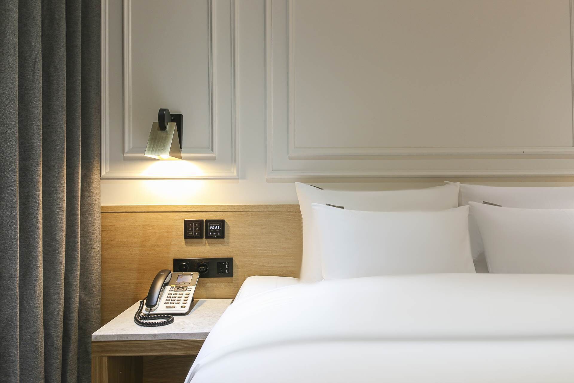 A hotel room with a bed, a bedside table with a phone, a wall-mounted light, and a control panel.