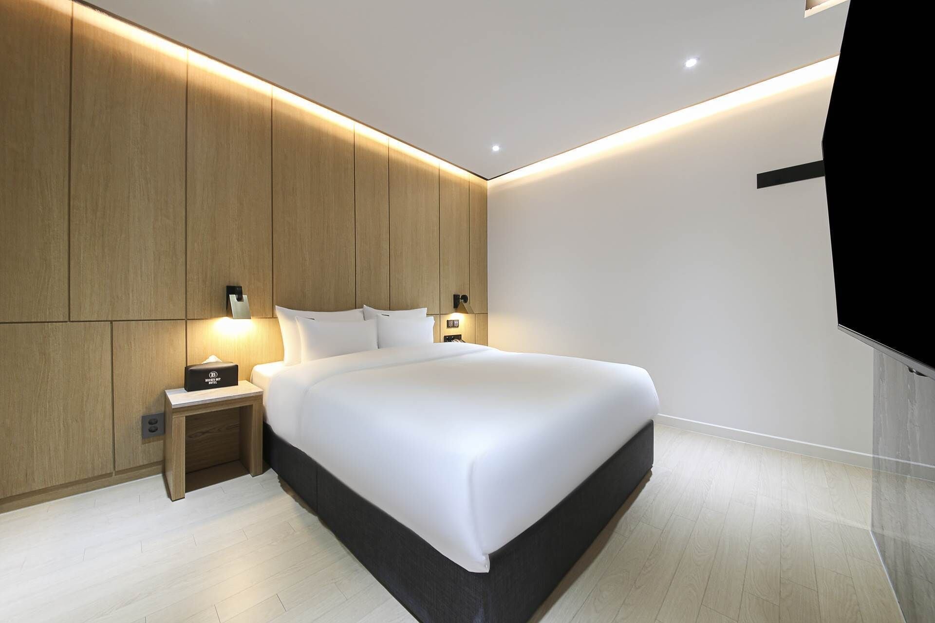 A modern hotel room with a large bed, wooden paneling on the walls, a bedside table with a lamp, and a flat-screen TV mounted on the wall.