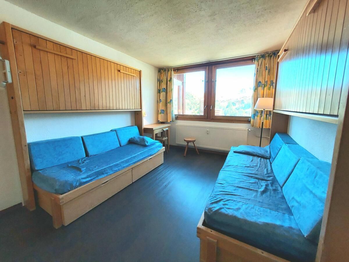 Ein kleines Zimmer mit zwei Holzbänken und einem Tisch, einem Fenster mit Vorhängen und einem Heizkörper.