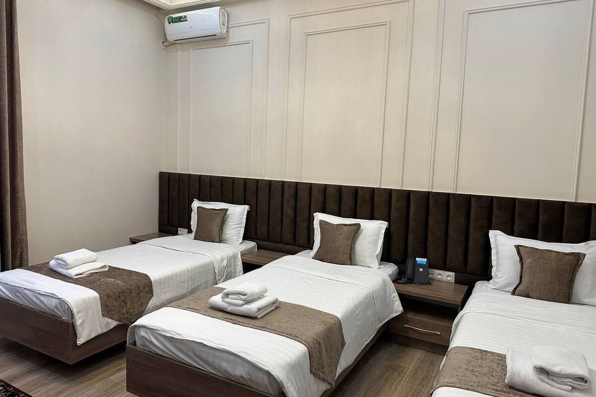 Habitación de hotel con tres camas individuales, cada una con cabecero marrón, ropa de cama blanca y una toalla doblada sobre la cama. Hay una mesita de noche con un teléfono y una botella de agua.