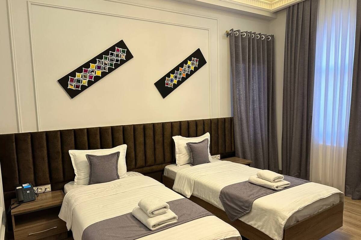 Habitación de hotel con dos camas, cabecera de madera, mesita de noche con teléfono y ventana con cortinas.