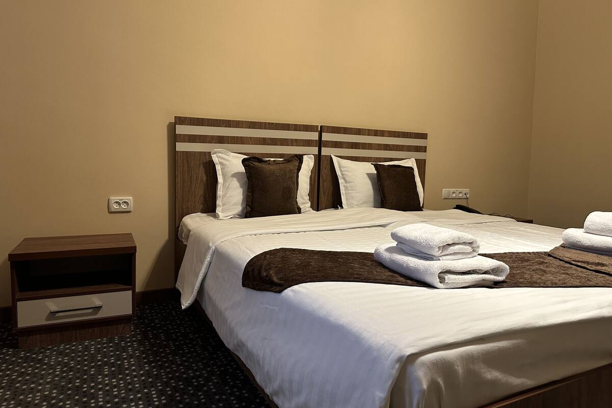 Una cama bien hecha con ropa de cama blanca y marrón, un cabecero de madera y una mesita de noche pequeña con un cajón.