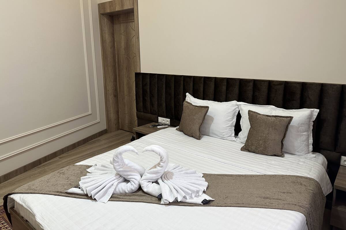 Una cama impecable con sábanas blancas y dos toallas con forma de cisne, un cabecero oscuro y una mesita de noche de madera con un control remoto.