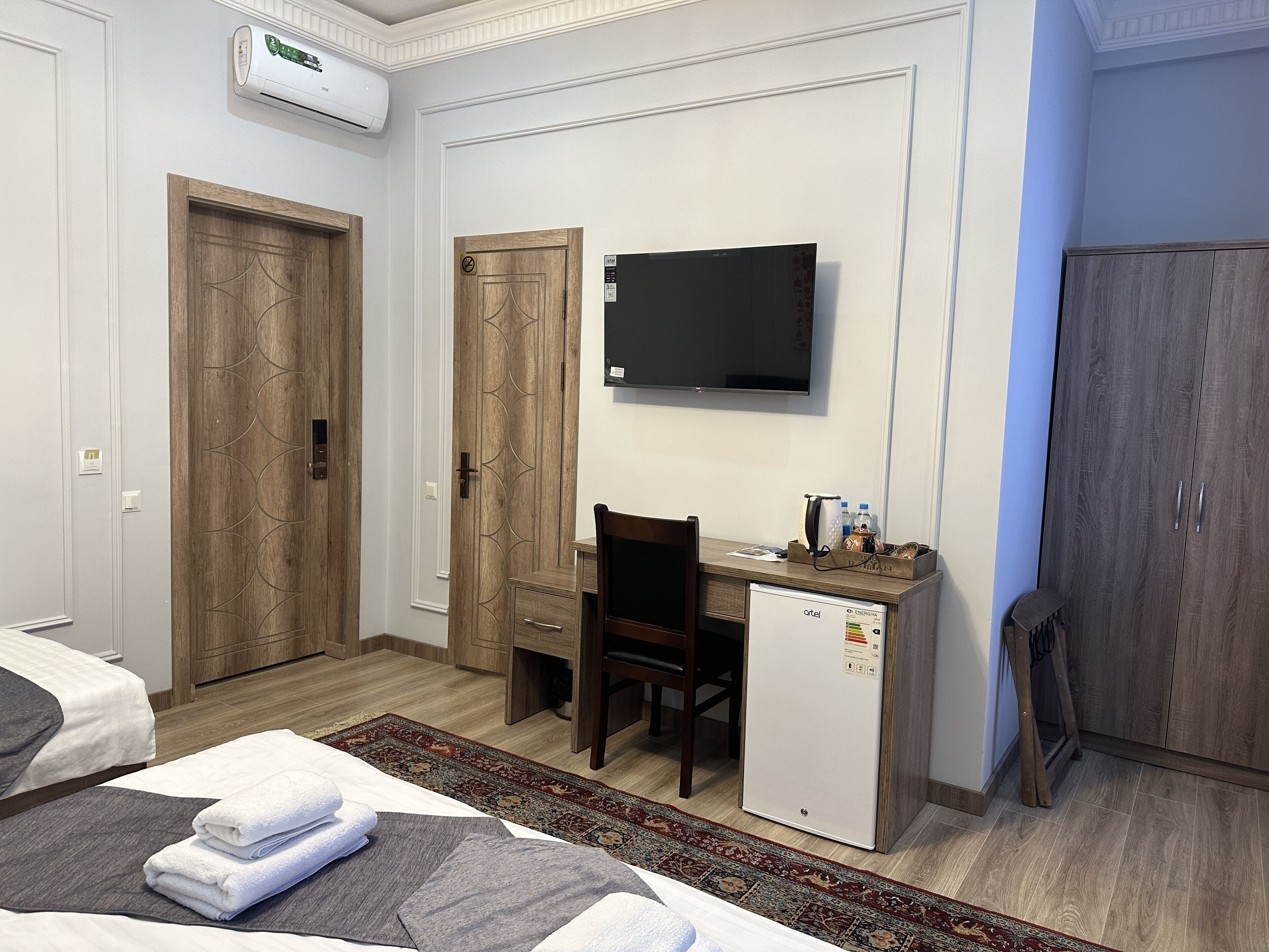 Ein Hotelzimmer mit zwei Holztüren, einem Schreibtisch mit Computer, einem Fernseher, einem Kleiderschrank und einem Bett mit Handtüchern.