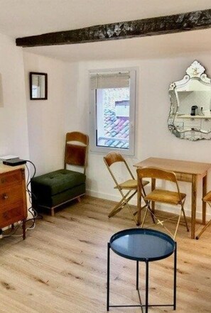 A room with a wooden floor, a small green sofa, a wooden table with chairs, a round black table, and a decorative mirror on the wall. - Petit Cocoon Aixois (Aix-en-Provence)