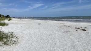 Une vaste plage de sable fin, avec des vagues douces, un ciel dégagé et des bâtiments qui se dessinent à l’horizon.