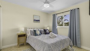 Une chambre à coucher comprenant un lit, une table de chevet, un ventilateur de plafond, une fenêtre avec des rideaux et une vue sur des palmiers.