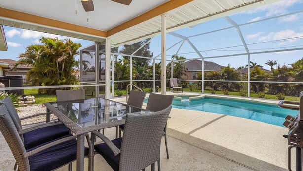 Une terrasse extérieure avec une table et des chaises en verre, un ventilateur de plafond et une vue sur une piscine et la végétation environnante.
