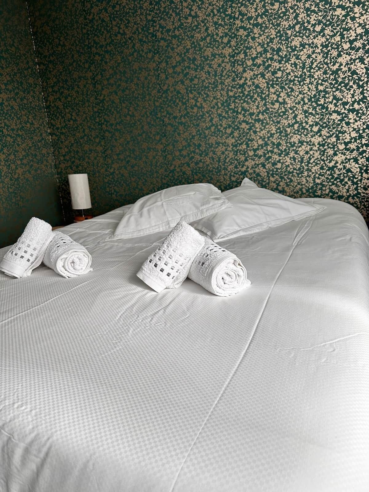 Una cama bien hecha con sábanas blancas y toallas enrolladas, un almohadón blanco y una pared con estampado floral.